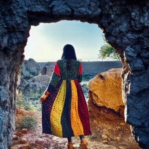 A portrait photograph of an Afghan girl, seen from behind, standing in the doorway of a stone structure. She wears a colorful dress and looks out towards a sunlit landscape, symbolizing hope and future prospects.