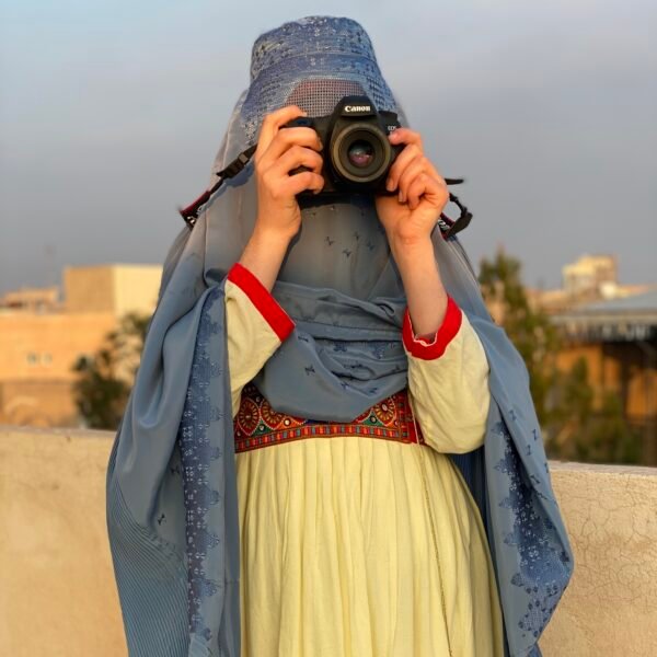 A portrait of an Afghan woman photographer, holding a Canon camera to her eye, her face partially obscured by the camera and her blue hijab. She is wearing a traditional Afghan dress.