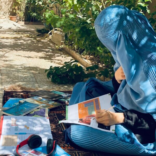 A portrait of an Afghan schoolgirl wearing a full blue hijab, sitting outdoors and studying her books. Red headphones are placed on the ground beside her. Emphasizes determination and perseverance in difficult conditions.