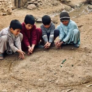 Four young Afghan boys are intently focused on playing a traditional game with small objects on a dusty ground circle in a street photography style image.