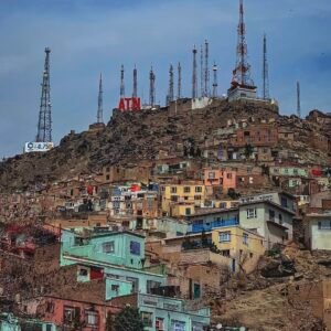 A panoramic street photography shot of colorful houses nestled on a mountainside in Kabul, Afghanistan, with communication towers silhouetted against the sky.