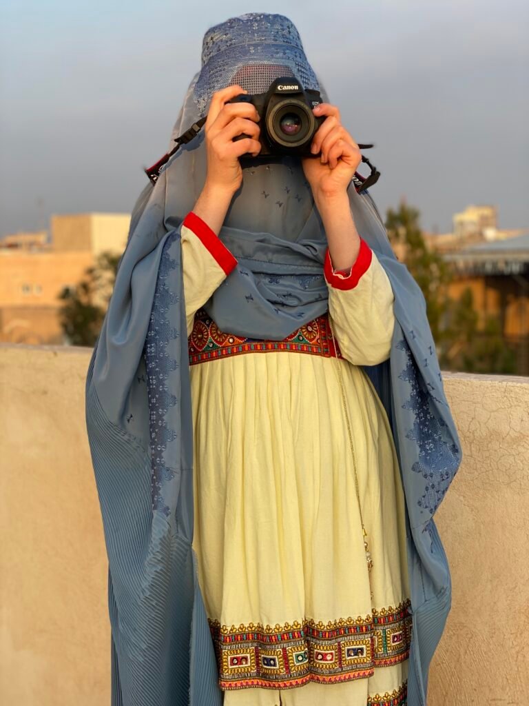 A portrait of an Afghan woman photographer, holding a Canon camera to her eye, her face partially obscured by the camera and her blue hijab. She is wearing a traditional Afghan dress.